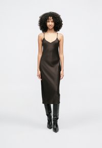 Sötétbarna selyemruha, karcsú szabással, V-nyakkivágással és vékony, állítható pántokkal, térdig érő fekete csizmával párosítva.