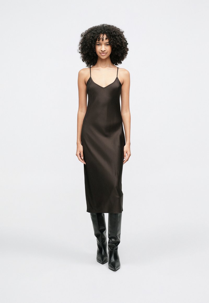 Sötétbarna selyemruha, karcsú szabással, V-nyakkivágással és vékony, állítható pántokkal, térdig érő fekete csizmával párosítva.