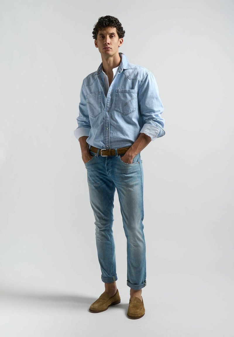 Chemise en denim bleu clair avec deux poches sur la poitrine, associée à un jean délavé et des chaussures de suède beige à enfiler, se tenant devant un fond neutre.