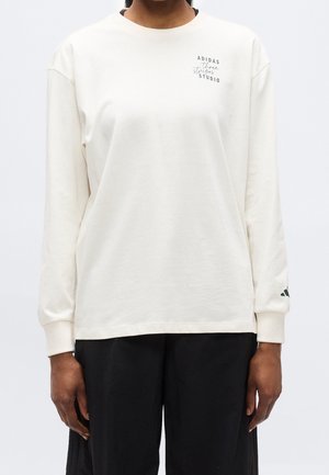Person iført en løs, langærmet hvid Adidas-sweatshirt med sort tekst på brystet og sorte bukser, står mod en ensfarvet baggrund.