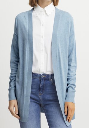 Personne portant un cardigan bleu clair ouvert sur une chemise blanche boutonnée à col festonné et un jean bleu en denim, mains détendues vers le bas.