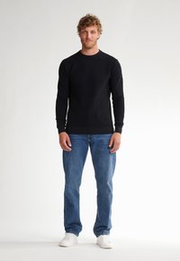 Black long-sleeve crewneck sweater with ribbed cuffs and hem, paired with light blue jeans and white sneakers, standing against a neutral background.