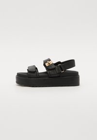 Black platform sandals with two adjustable straps, gold-tone hardware, and a textured sole. Made of smooth leather with a sleek design.