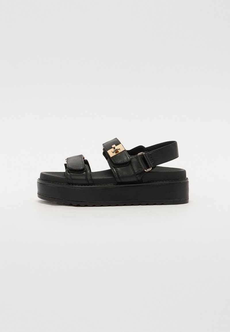 Black platform sandals with two adjustable straps, gold-tone hardware, and a textured sole. Made of smooth leather with a sleek design.