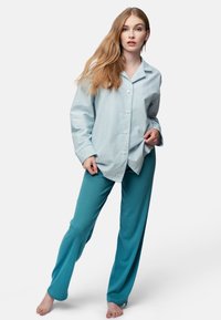 Camicia button-up a righe blu e bianche con maniche lunghe abbinata a pantaloni a gamba larga solidi in teal, a coste. Modella in piedi a piedi nudi.