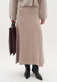 Šviesiai rudos spalvos megzta midi sijonas, turintis glotnią tekstūrą; derinamas su juodais smailiais bateliais su aukštu kulnu ir tamsiai rudą rankinę, laikoma rankoje.