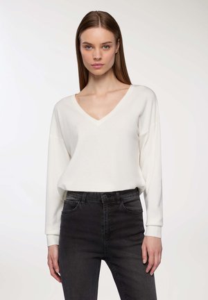 Maglione crema a V realizzato in tessuto morbido, con maniche lunghe, abbinato a jeans neri a vita alta con tasche frontali e silhouette aderente.