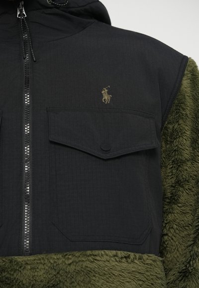 Casaco com capuz em preto e verde oliva, apresentando fecho de correr à frente, textura canelada, dois bolsos frontais e um logótipo no peito. Material suave e peludo.