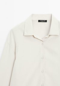 Light beige button-up shirt with a collar, featuring a smooth texture and an A-line shape. It has long sleeves and a subtle sheen.