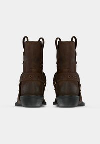 Ζευγάρι καφέ δερμάτινων μποτών αστραγάλου με λουριά και θηλιές, εμφανίζονται από πίσω σε απλό λευκό φόντο.