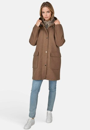 Junge Frau, die einen braunen Wintermantel mit Kapuze und großen Taschen trägt, hellblaue Jeans und weiße Sneakers, steht vor einem schlichten Hintergrund.