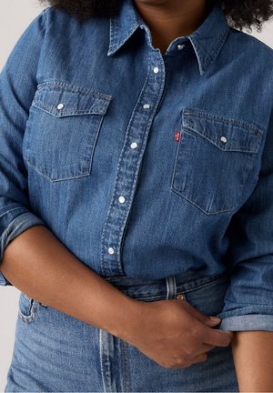 Personne portant une chemise en denim bleu avec des boutons-pression nacrés et des poches poitrine, remontant une manche, associée à un jean bleu en denim.