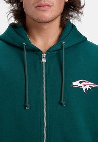 Zelená mikina na zips vyrobená z jemného materiálu, s tkaničkami, strieborným zipsom a grafickým logom v bielej a červenej farbe na hrudi.
