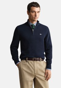 Marineblauwe trui met ritssluiting en geribbelde manchetten, gedragen over een gestreept overhemd. Gecombineerd met lichtkaki broek en een bruine riem. Voorzien van logodetails.