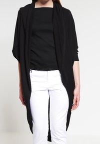 Svart öppen cardigan med långa ärmar, tillverkad av mjukt tyg, med en avslappnad passform och flödande design, bärs över en svart topp och vita byxor.