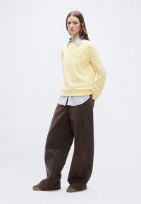 Νέα γυναίκα στέκεται με τα χέρια στις τσέπες, φοράει κίτρινο φούτερ πάνω από ριγέ πουκάμισο και φαρδιά καφέ παντελόνια με αθλητικά παπούτσια.