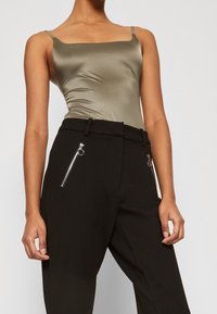 Olive zelený saténový body s tenkými ramínky, spárovaný s černými kalhotami s vysokým pasem, které mají stříbrné kapsy na zip. Hladká textura, přiléhavý střih.