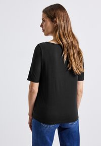 Femme aux longs cheveux bruns portant un haut noir à manches courtes et un jean bleu, se tenant de profil devant un fond blanc uni.