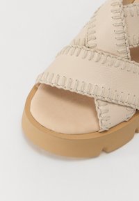 Sandalo in pelle beige con cinturini incrociati, dettagli cuciti, soletta morbida e una spessa suola in gomma marrone chiaro.