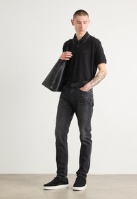 Schwarzes Poloshirt mit weißem Kragen, dunkle Jeans, schwarze Sneaker und eine schwarze Ledertasche. Auf dem Arm ist ein sichtbares Tattoo zu sehen.