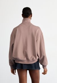 Sweatshirt oversized, em cor malva, com colarinho alto e ajuste largo. Tecido liso e punhos canelados; usado com uma saia escura e em camadas.