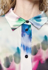 Blouse à col avec des boutons noirs et un motif aquarelle aux teintes bleues, vertes, roses et grises sur un tissu soyeux.
