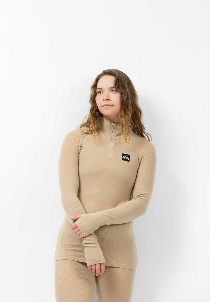 Haut beige à manches longues et à zip, en tissu côtelé. Présente des trous pour les pouces aux poignets et une petite patch logo sur la poitrine.
