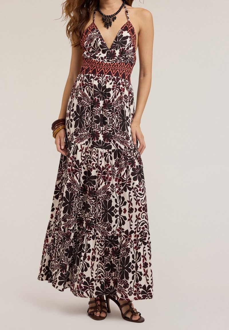 Femme portant une robe fleurie sans manches, longue jusqu'au sol, avec un profond décolleté en V et des talons noirs à brides, accessoirisée avec des bracelets et un collier.