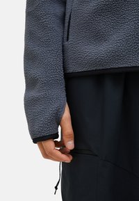 Gray fleece jacket with a textured surface, black trim, and a side zipper pocket. Hand grasping the pocket zipper is visible.