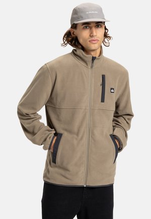 Tan fleece jacket with a zippered chest pocket and black accents. Features a high collar and elastic cuffs. Worn with dark pants.