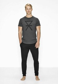 Grå kortærmet t-shirt med et sort stribet grafisk design på forsiden, parret med sorte slim-fit bukser; barfodet.