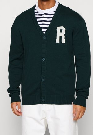Homme portant un cardigan boutonné vert foncé avec un grand "R" blanc sur la poitrine, par-dessus une chemise rayée blanche et marine, assorti à un pantalon blanc.
