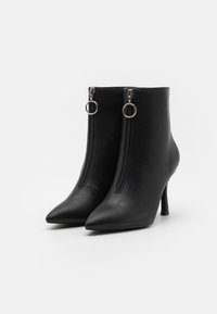 Bottes noires en cuir à la cheville avec bouts pointus, fermetures éclair à l'avant munies de tirettes en anneau argenté, et talons aiguilles sur fond blanc.