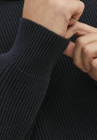 Polsino scuro a coste con un design aderente, che mostra un tessuto a trama e una finitura pulita. È visibile una mano che regola la manica.