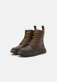 Botas de cuero marrón con cordones amarillos, suela de goma negra y costuras amarillas. Cuentan con una pestaña en la parte trasera para facilitar su uso.