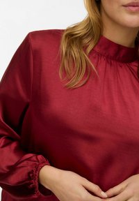 Bordeaux satijnen blouse met een hoge hals en geplooide details, voorzien van lange mouwen met elastische boorden en een glad, glanzend oppervlak.