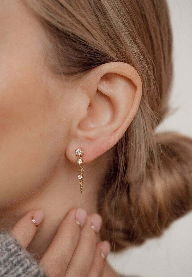 Orecchini d'oro con un design a catena che presenta tre piccoli gemme trasparenti, appesi elegantemente vicino all'orecchio, arricchiti da uno sfondo morbido e neutro.