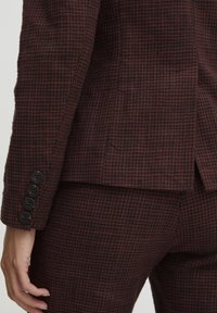 Blazer à carreaux sombres en tissu texturé. Présente une coupe ajustée, quatre boutons noirs sur les poignets et une fente à l'arrière.