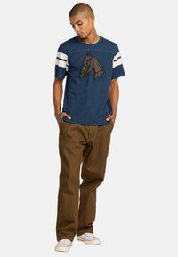 T-shirt en coton marine à manches courtes, avec des rayures blanches et un graphique de cheval sur le devant ; associé à un pantalon marron et des baskets blanches.