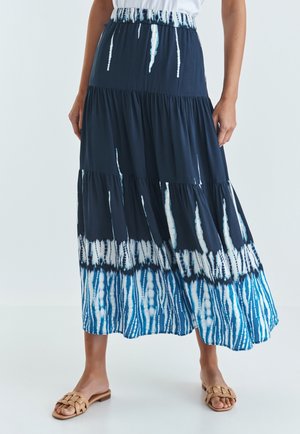 Kobieta w granatowo-białej falującej, warstwowej spódnicy maxi tie-dye oraz beżowych plecionych płaskich sandałach, stojąca na białej podłodze.