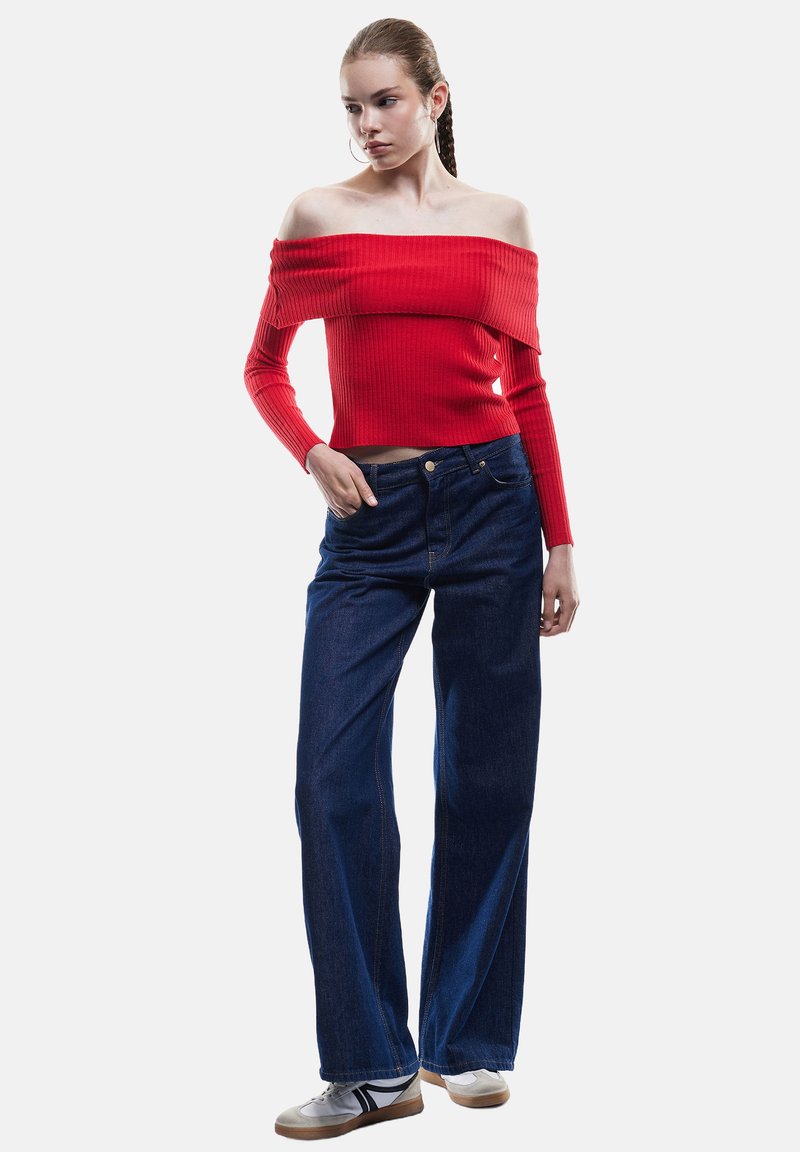 Czerwony sweter z dekoltem typu off-the-shoulder i ribbrowanym wzorem, zestawiony z szerokimi niebieskimi dżinsami i białymi trampkami, o luźnym kroju i teksturowanym materiale.