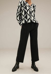 Blusa geometrica in bianco e nero con apertura frontale a bottoni, abbinata a pantaloni neri a gamba larga. Modella indossa stivaletti neri fino alla caviglia.