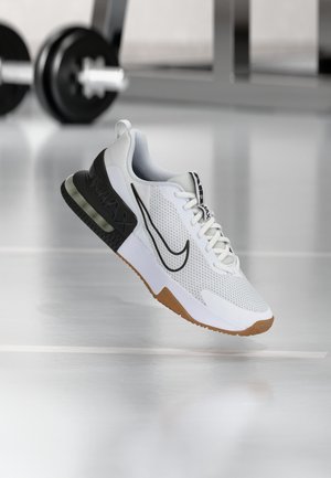 Chaussure de sport Nike Air Max blanche et noire avec tige en mesh, semelle en gomme et coussin d'air visible flottant au-dessus d'un sol de salle de sport avec des poids en arrière-plan.
