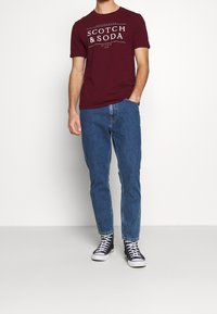 Burgunder kortærmet T-shirt med hvid tekst, parret med blå denim jeans og sorte høy-top sneakers med hvide såler og snørebånd.