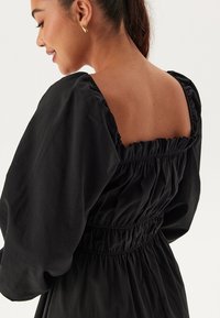 Zwarte jurk met opgezette mouwen, elastische taille, vierkante achterkant en geplooide textuur. De gladde stof biedt een zachte afwerking met een ontspannen pasvorm.