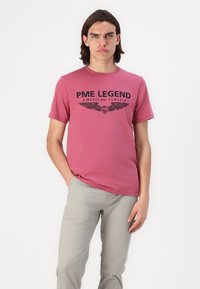 T-shirt bordeaux avec un logo noir imprimé "PME LEGEND" et des graphiques d'ailes. Porté avec un pantalon gris clair, avec des manches courtes et un design col rond.