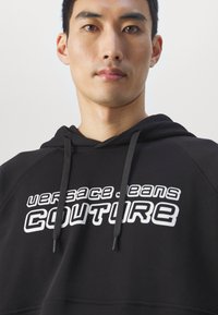 Black hoodie with a kangaroo pocket, featuring a white graphic logo “VERSACE JEANS COUTURE” in bold, stylized font on the chest.