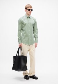 Homem vestindo uma camisa de botão verde menta, calças creme, sapatos pretos e óculos de sol, segurando uma grande bolsa tote de couro preta.