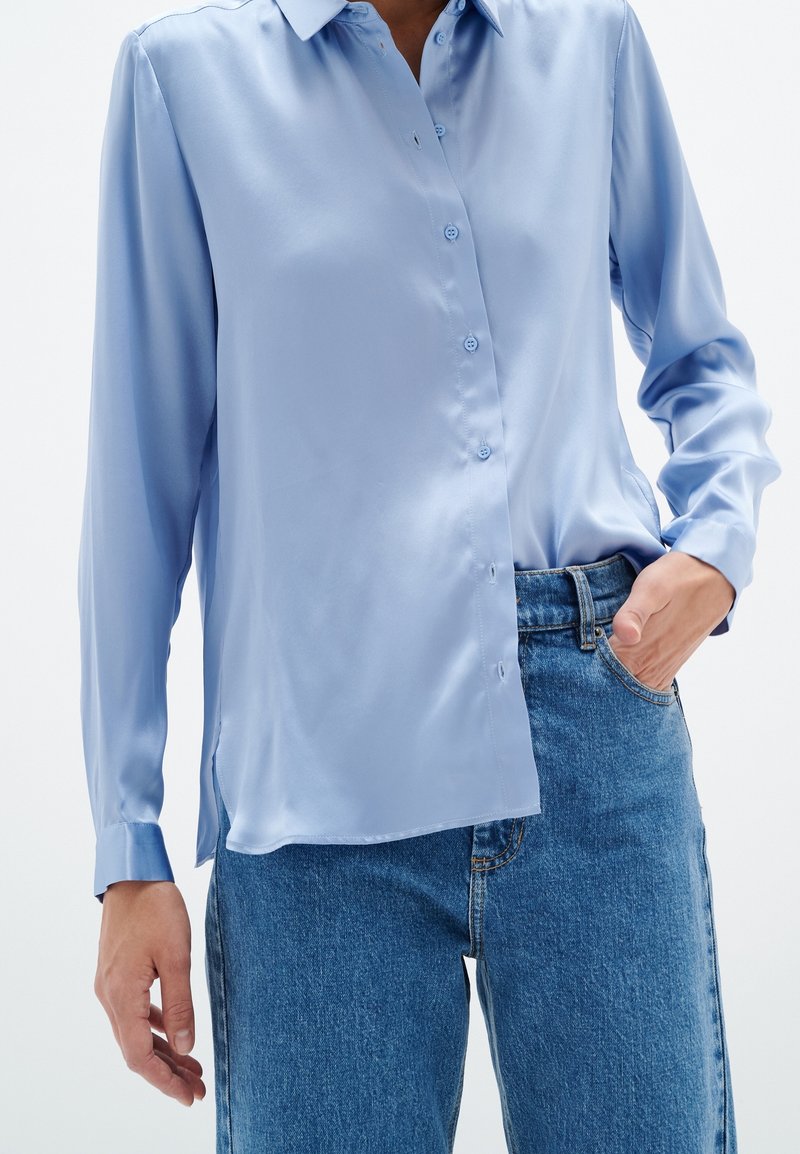 Camisa de satén azul claro con mangas largas, cierre de botones en la parte delantera y un dobladillo ligeramente curvado, combinada con vaqueros de talle alto.
