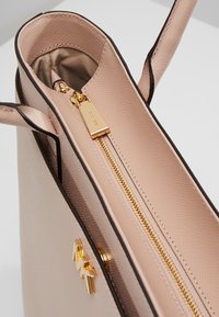 Sac à main en cuir beige texturé avec fermeture éclair dorée et logo, ouverture sur le dessus montrant les compartiments intérieurs et double poignée supérieure.